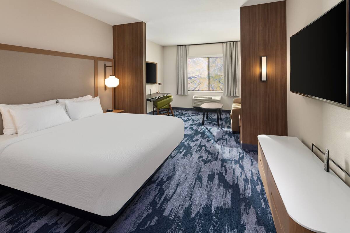 A hotel room with a large bed, a desk with a chair, a TV, and a large window with curtains.