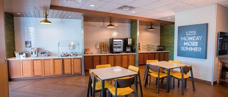 A modern cafe with wooden flooring, yellow chairs, a white table, and a kitchen area with a vending machine.