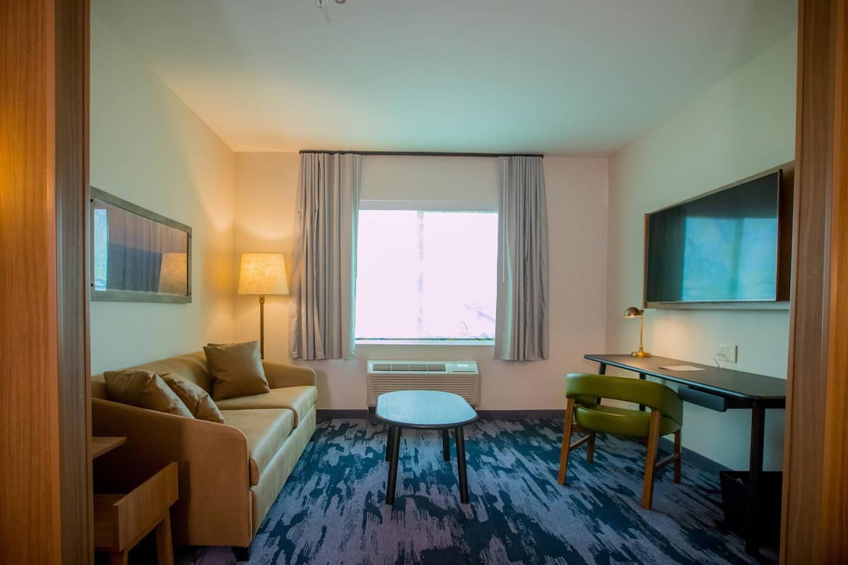 A hotel room with a sofa, a small table, a chair, a desk, a lamp, a TV, and a window with curtains.