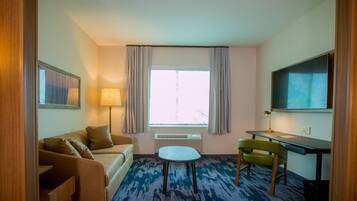 A hotel room with a sofa, a small table, a chair, a desk, a lamp, a TV, and a window with curtains.