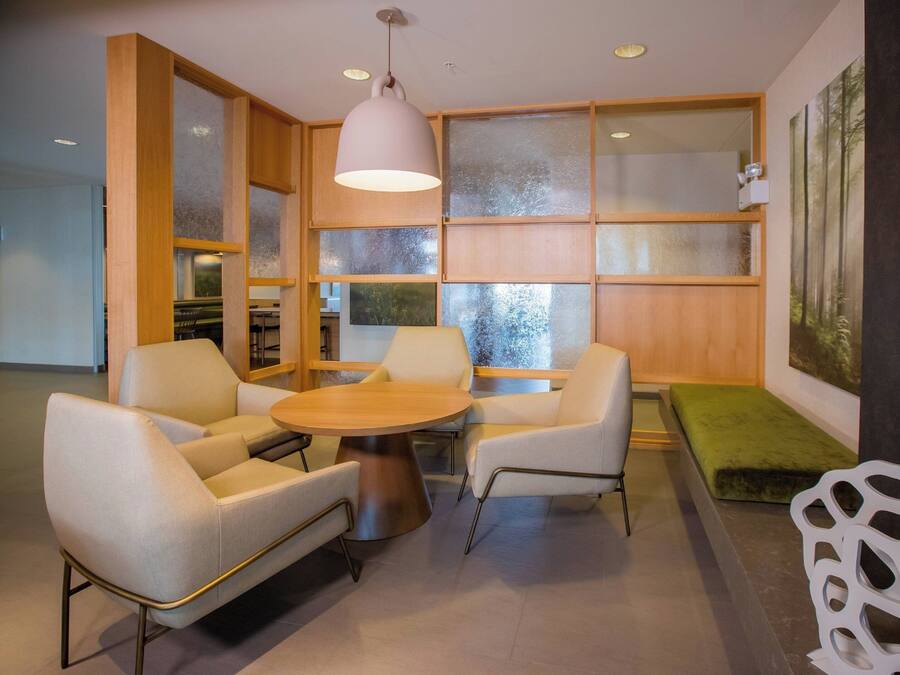A modern interior with a round table, two white armchairs, a green bench, and a large window with frosted glass panels.