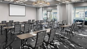 Un salón de conferencias con varias mesas y sillas, una pantalla grande y vistas a edificios a través de amplias ventanas.
