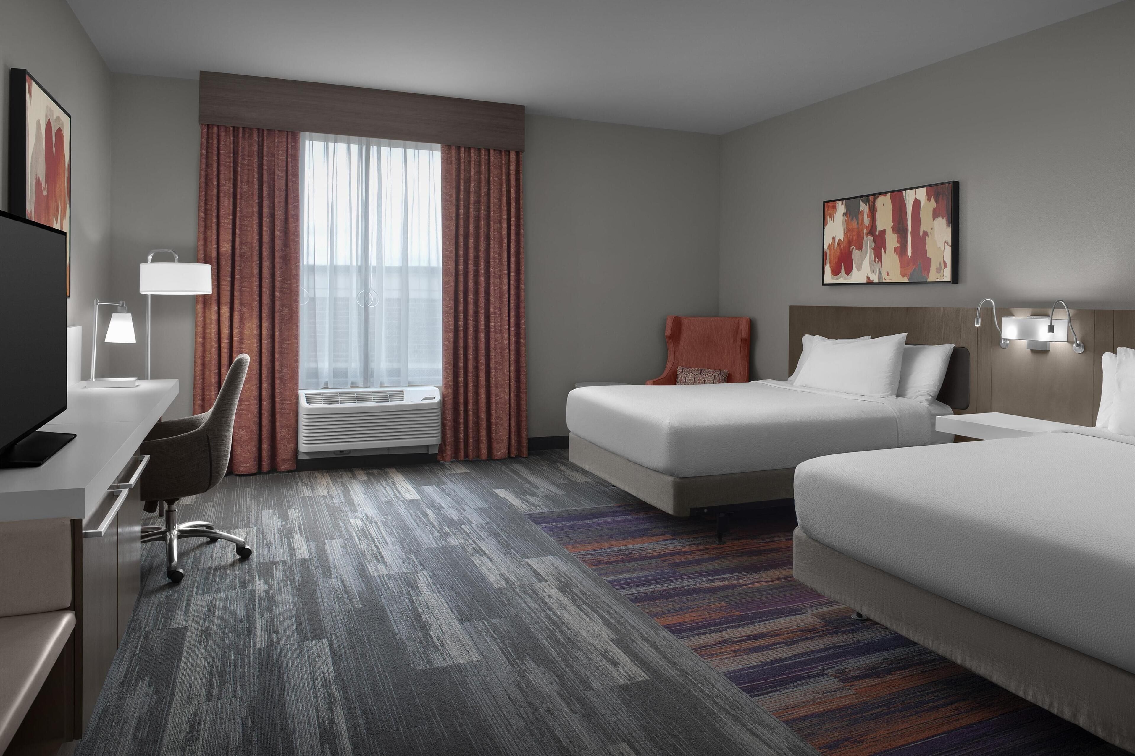 A hotel room with two beds, a desk with a chair, a TV, and a large window with curtains.