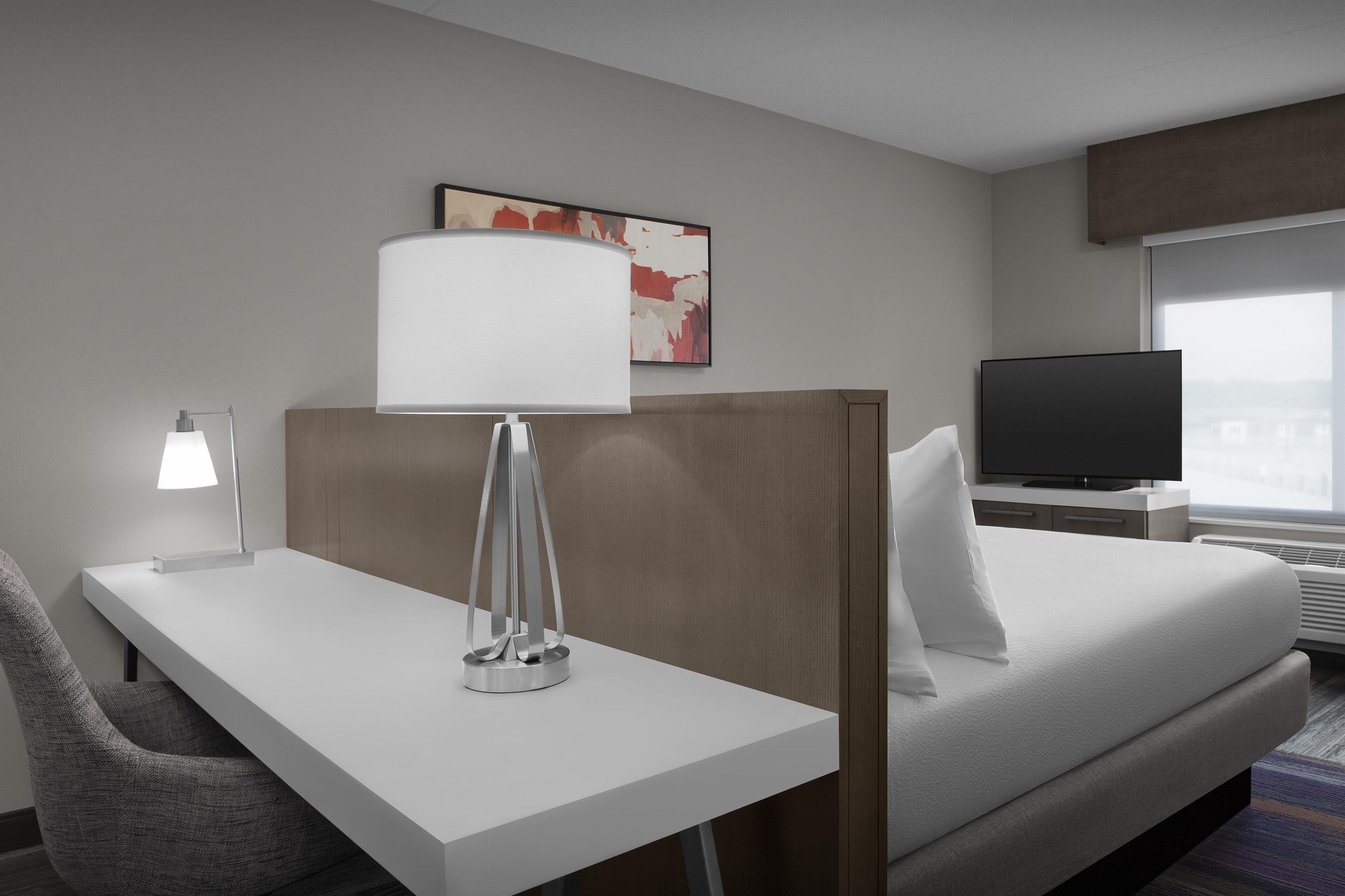 A hotel room with a bed, a desk with a lamp, a chair, a television, and a window.