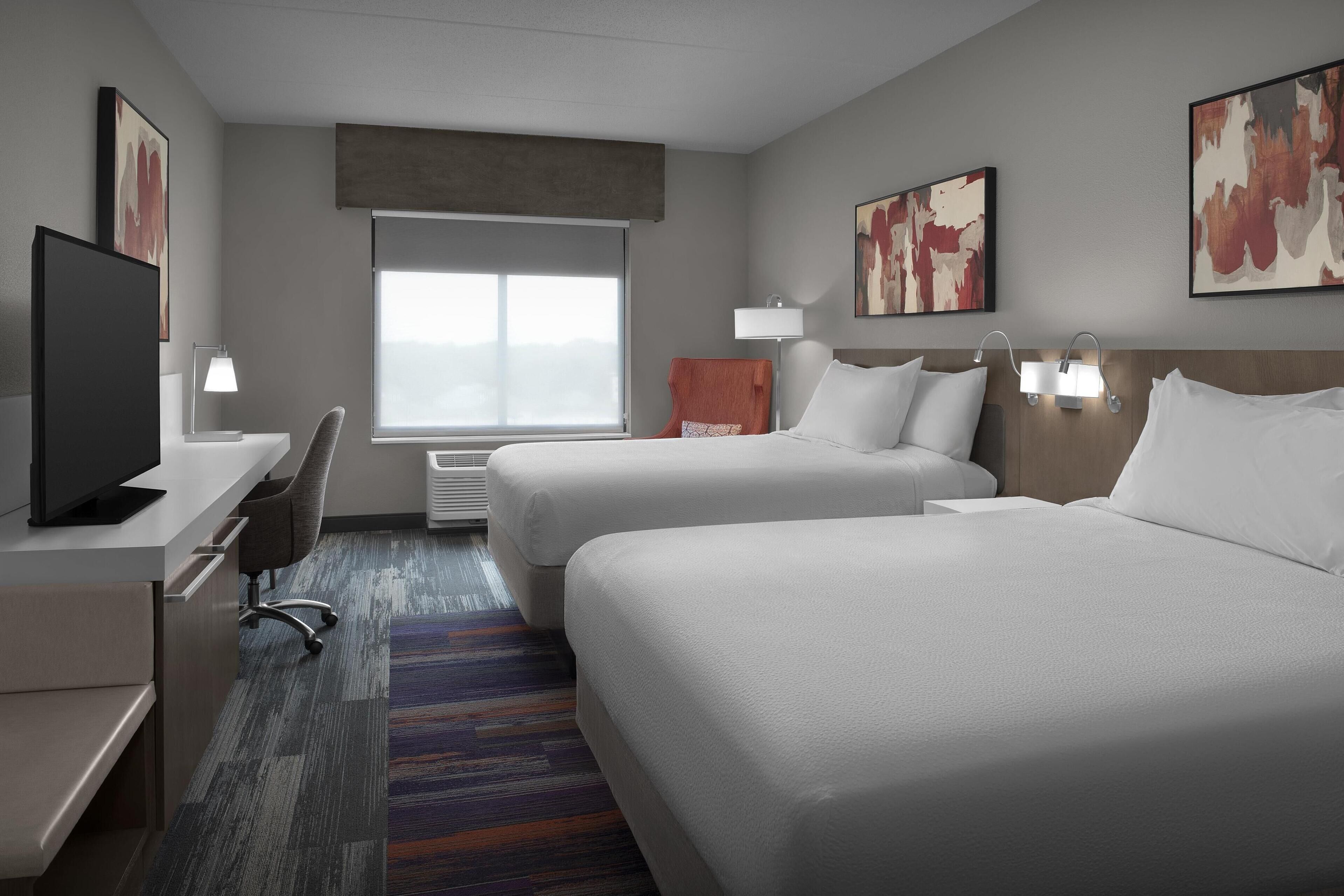 A hotel room with two beds, a desk, a TV, and a large window.