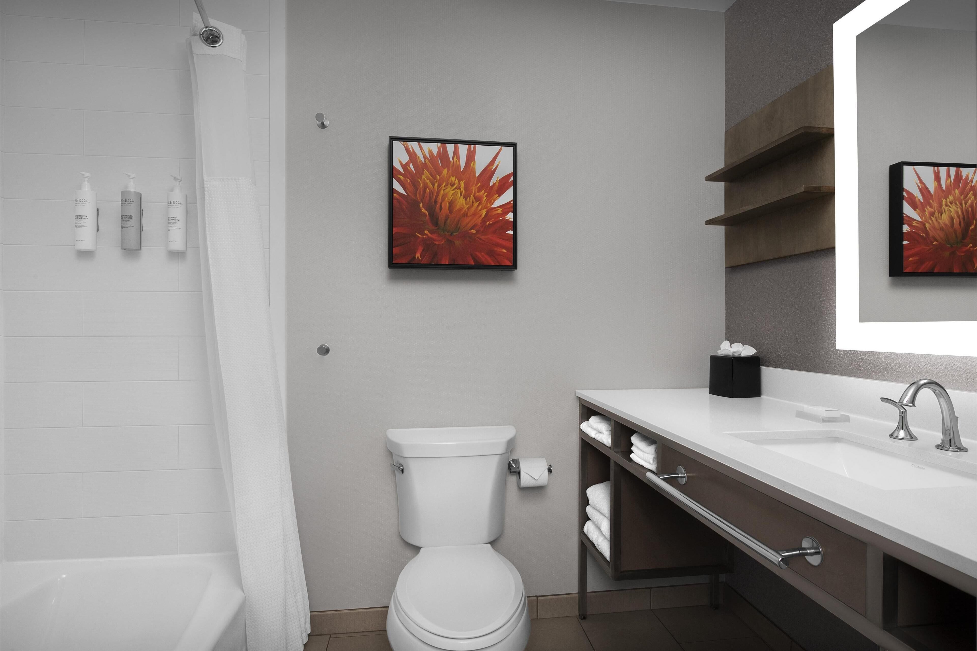 A modern bathroom with a toilet, sink, and shower. There are three toiletry bottles on the wall and a framed picture above the toilet.