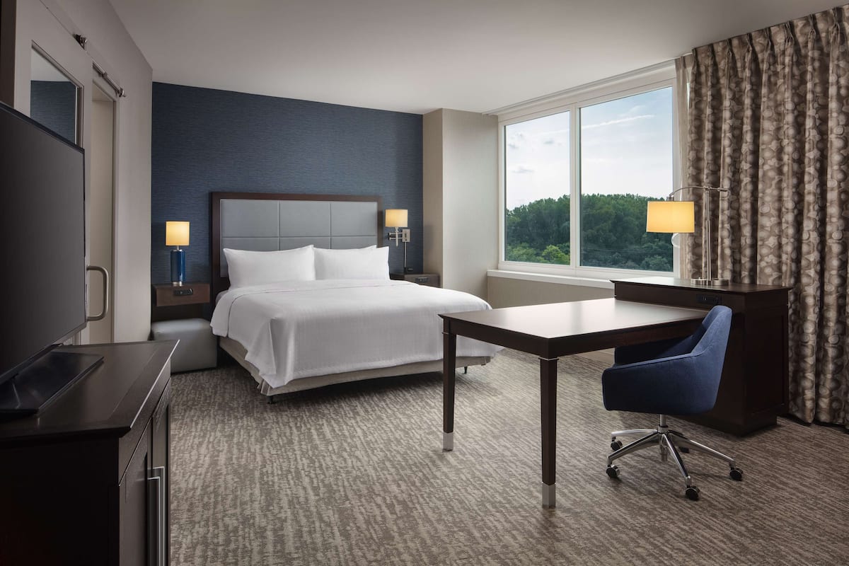 Ein Hotelzimmer mit Bett, Schreibtisch, Stuhl und großem Fenster mit Vorhängen.