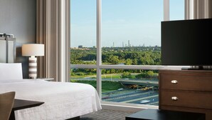 A hotel room with a large bed, a TV, a desk, and a view of a cityscape through large windows. - Homewood Suites by Hilton Teaneck Glenpointe (Teaneck)