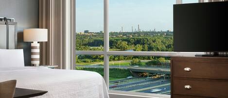 Habitación de hotel con una cama grande, televisión, escritorio y vistas a un paisaje urbano a través de amplios ventanales.