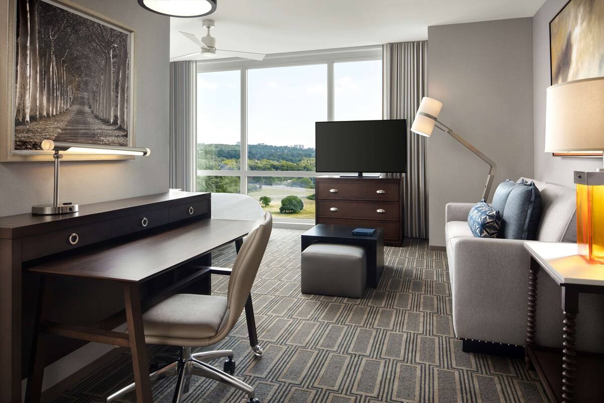 Ein Hotelzimmer mit Schreibtisch, Stuhl, Fernseher, Sofa und einem großen Fenster mit Blick ins Freie.