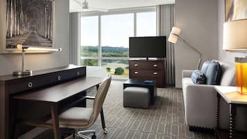 A hotel room with a desk, chair, TV, sofa, and a large window offering a view of the outdoors.