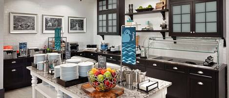 A modern kitchen with dark cabinetry, a marble countertop, and a central island with a fruit bowl. There are framed pictures on the wall, a chalkboard menu, and a glass display case.