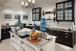 Una cocina moderna con armarios oscuros, encimera de mármol y una isla central con frutero. Se observan cuadros enmarcados en la pared, un menú en una pizarra y una vitrina de cristal.