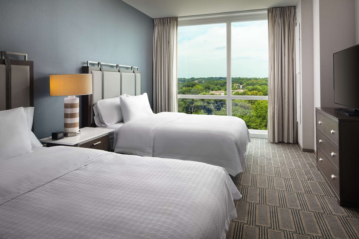 Ein Hotelzimmer mit zwei Betten, einem Fernseher, einer Lampe und einem großen Fenster mit Blick auf Grünflächen.