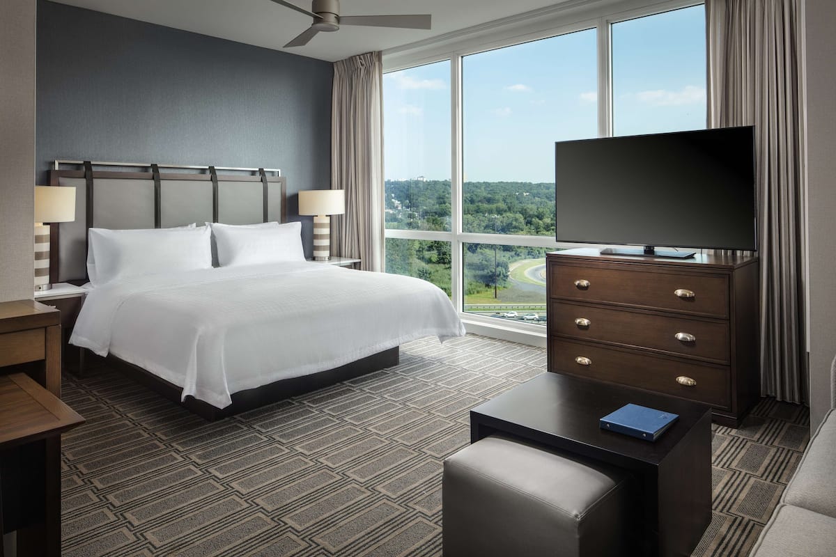 Ein Hotelzimmer mit einem großen Bett, einem Fernseher auf einer Kommode, einem Schreibtisch und Blick auf eine Stadtlandschaft durch große Fenster.