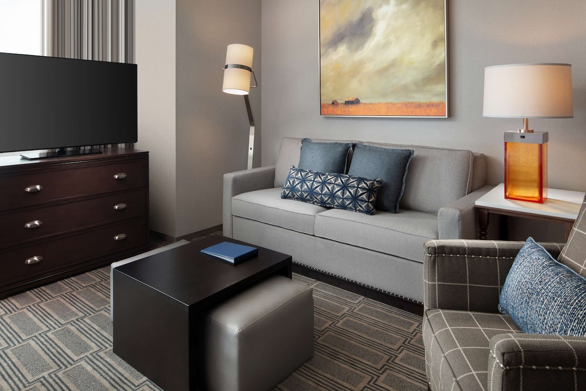 Ein modernes Hotelzimmer mit Flachbildfernseher, einer Couch mit gemusterten Kissen, einem Couchtisch, einer Lampe und einem Bild an der Wand.