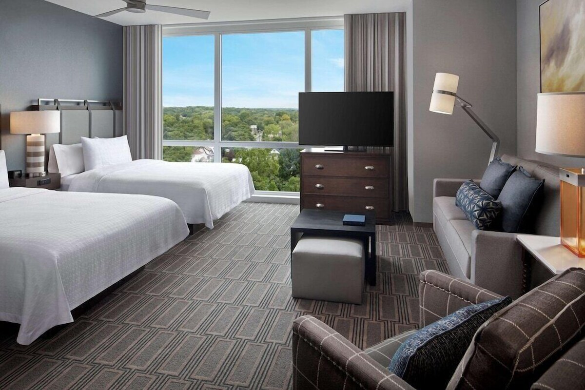 Habitación de hotel con dos camas, televisión, sofá, escritorio y ventana grande con vistas a la ciudad.