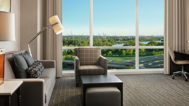 A hotel room with a large window offering a cityscape view, a sofa, a chair, a desk, and a lamp. - Homewood Suites by Hilton Teaneck Glenpointe (Teaneck)