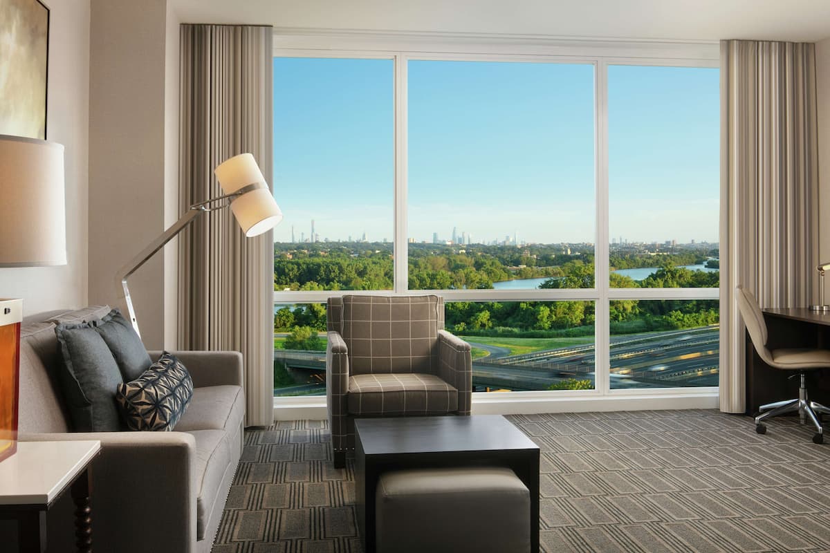 Ein Hotelzimmer mit großem Fenster und Blick auf die Stadt, inklusive Sofa, Sessel, Schreibtisch und Lampe.