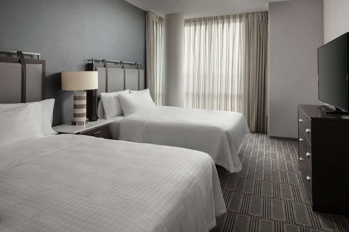 Ein Hotelzimmer mit zwei Betten, einem Fernseher, einer Lampe und einem großen Fenster mit Vorhängen.