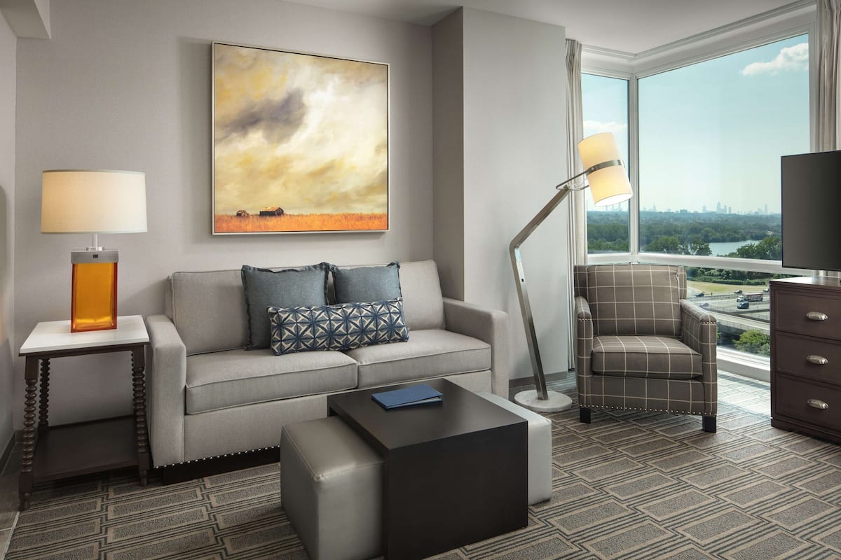 Ein modernes Hotelzimmer mit Sofa, Ohrensessel und Hocker, einem großen Fenster mit Blick auf die Stadt und einem Bild an der Wand.