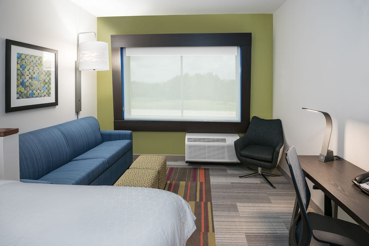 A hotel room with a bed, a blue sofa, a desk with a chair, a large window, and an air conditioner.