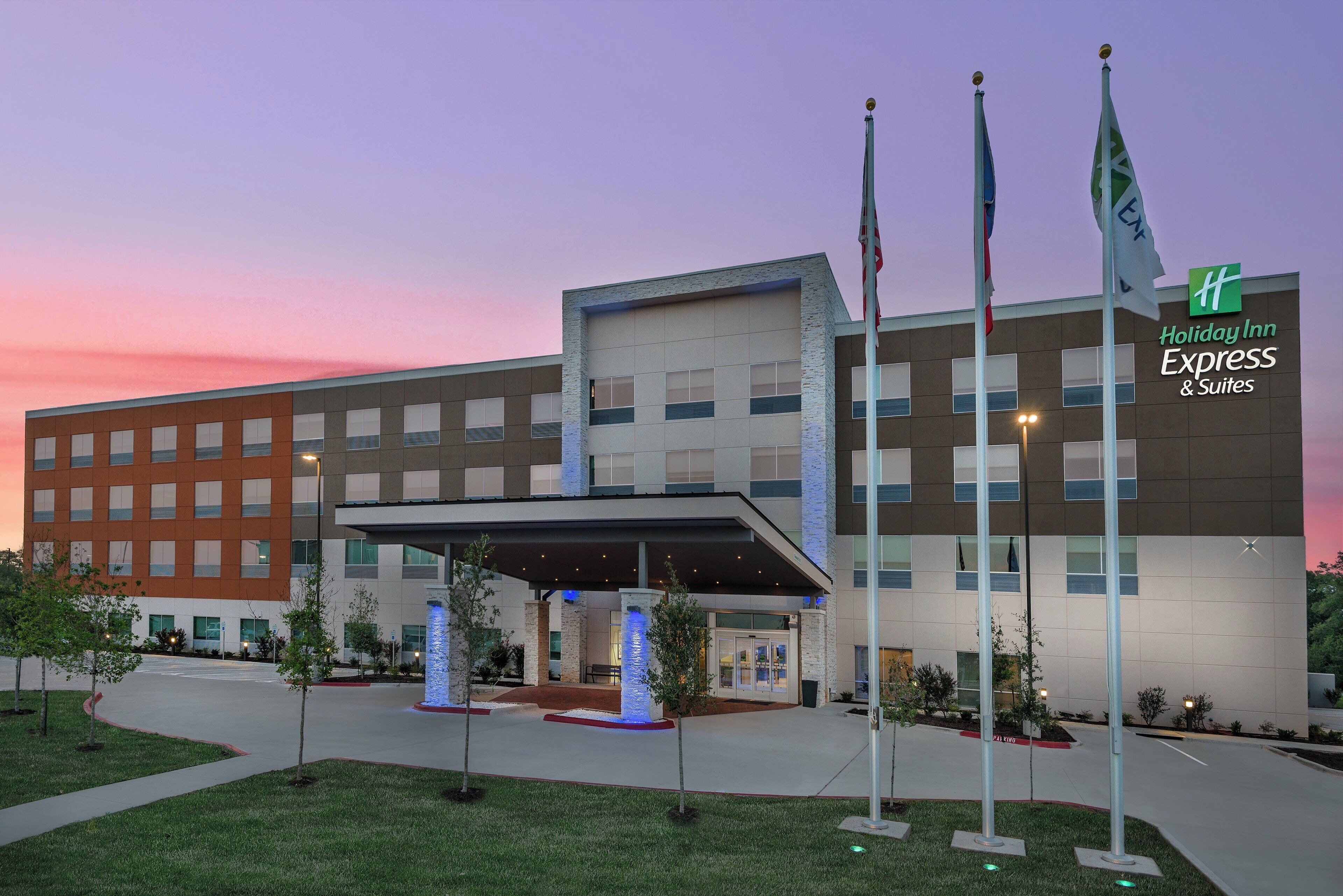 Edificio de Holiday Inn Express & Suites con entrada cubierta, varias banderas y un área de entrada ajardinada.