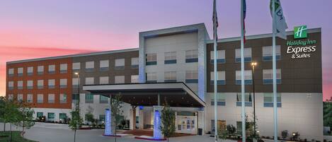Edificio de Holiday Inn Express & Suites con entrada cubierta, varias banderas y un área de entrada ajardinada.