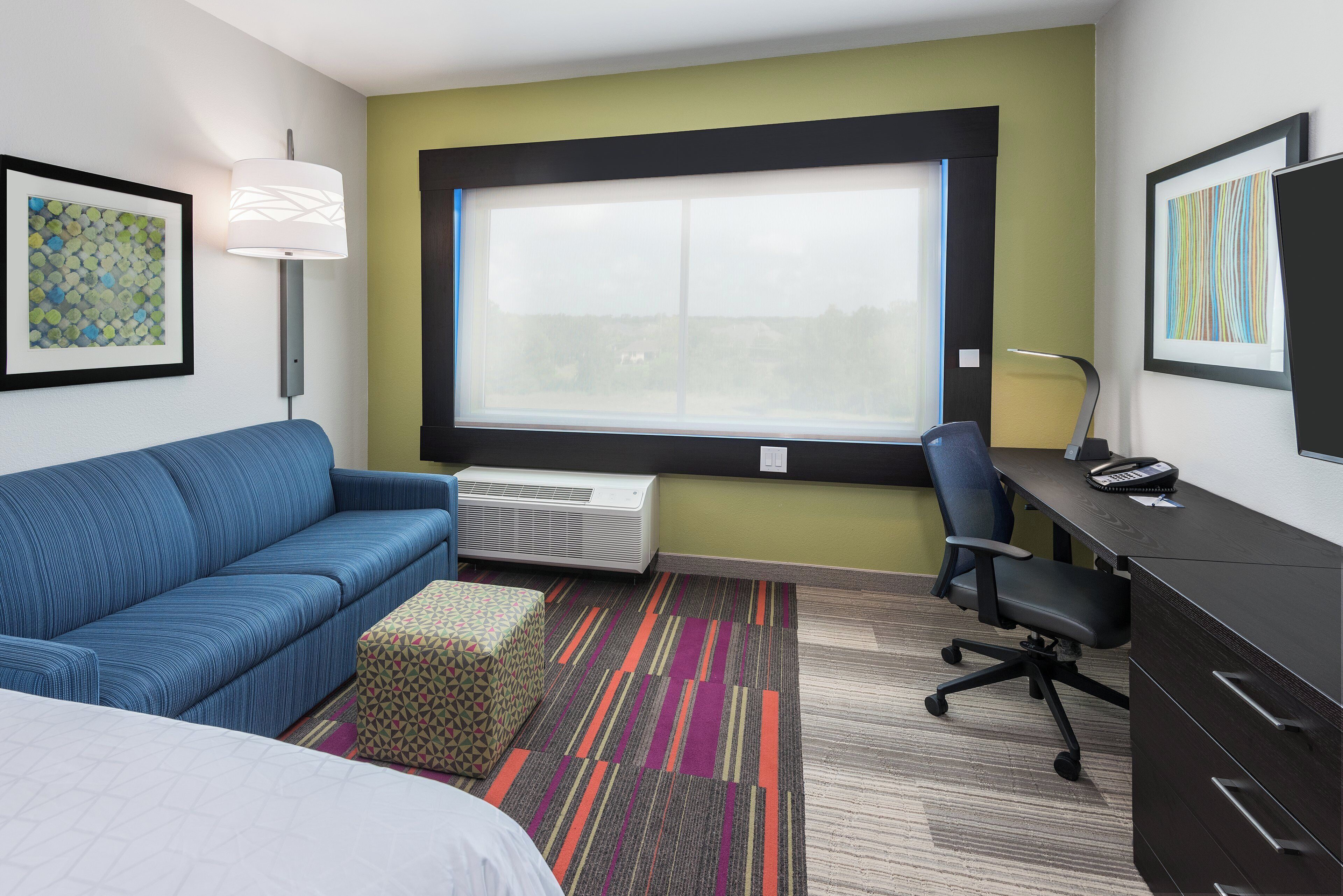 Une chambre d’hôtel avec un lit, un canapé bleu, un bureau avec une chaise, une fenêtre et des œuvres d’art accrochées aux murs.