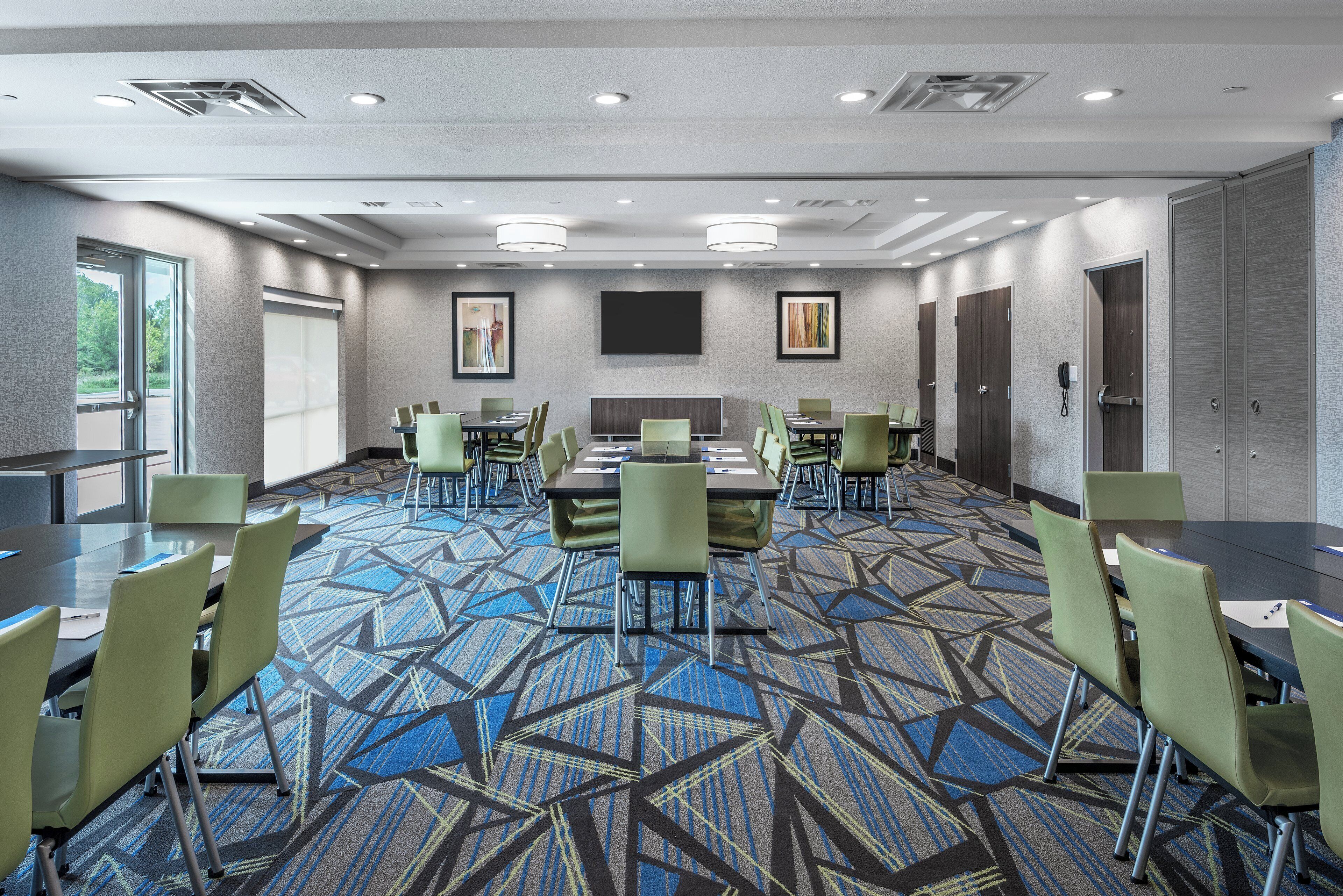 A conference room with a large table, multiple chairs, a flat-screen TV, and a patterned carpet.