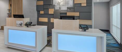 A hotel reception area with two reception desks, a wall-mounted information board, and a carpeted floor.