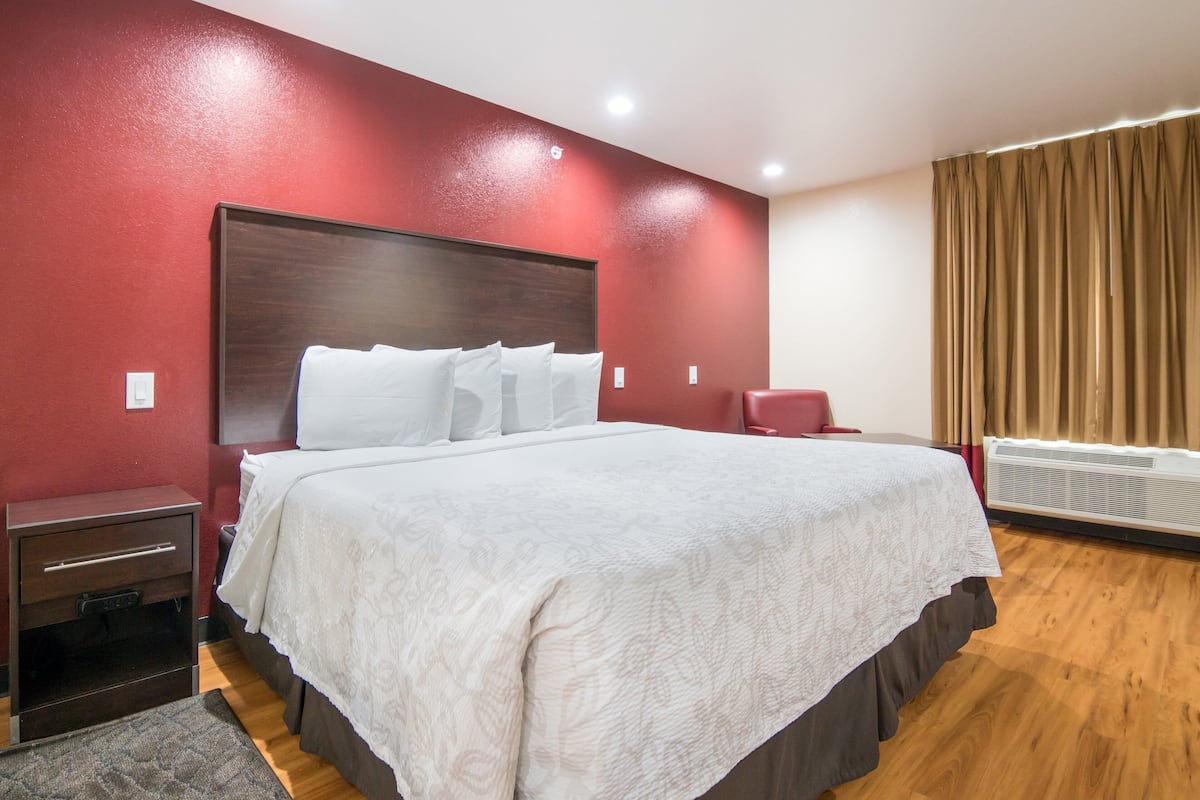 A hotel room with a large bed, a wooden headboard, a nightstand, a red chair, and a window with curtains.