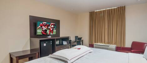 A hotel room with a bed, a television mounted on the wall, a desk, a chair, and a window with curtains.
