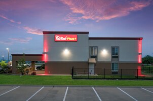 A Red Roof Plus building with a flat roof, a parking lot in front, and a clear sky.