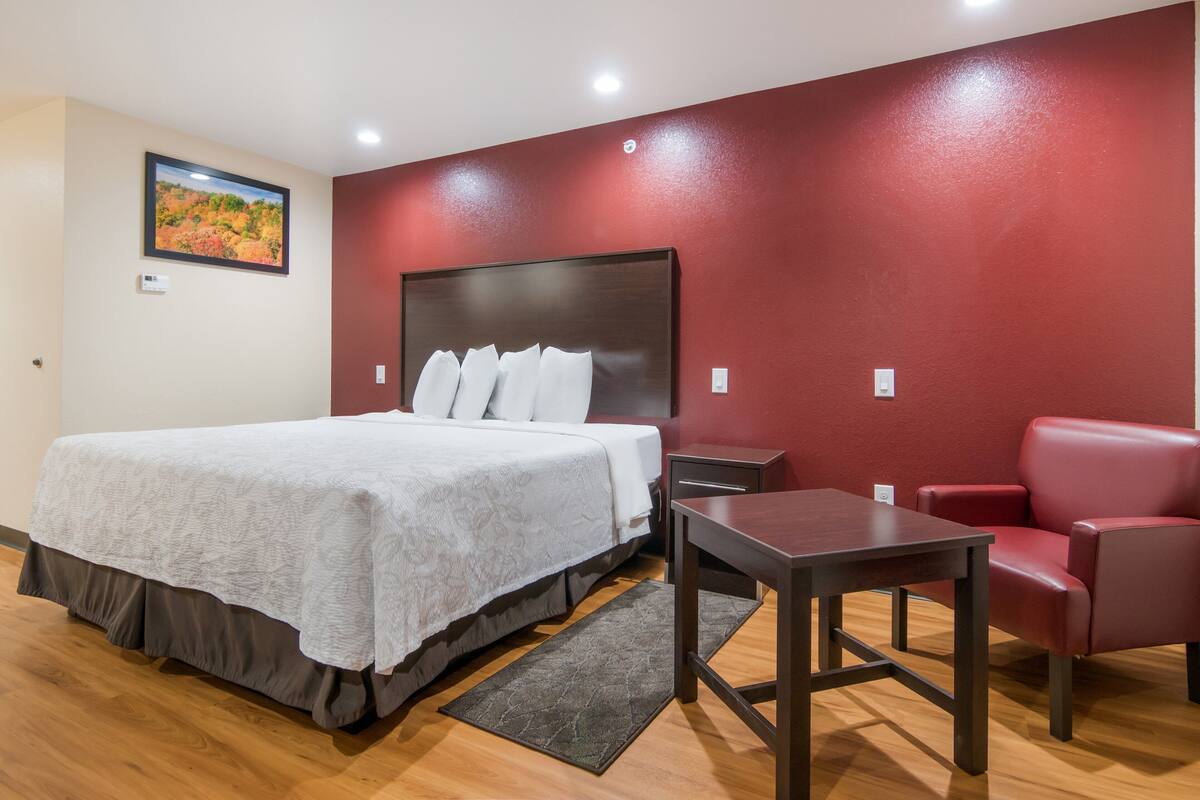 A hotel room with a large bed, a red wall, a wooden floor, a desk, and a red chair.
