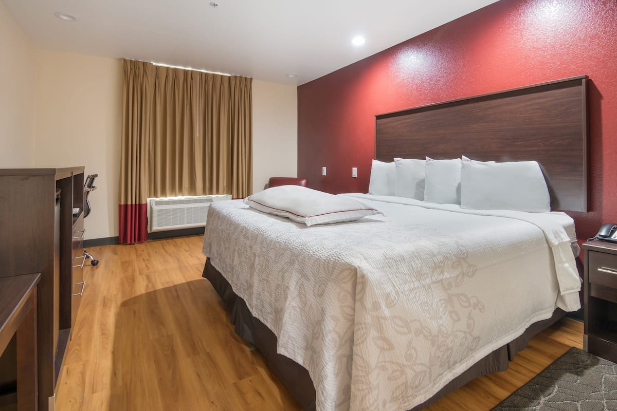 A hotel room with a large bed, a wooden floor, a red accent wall, and a window with curtains.