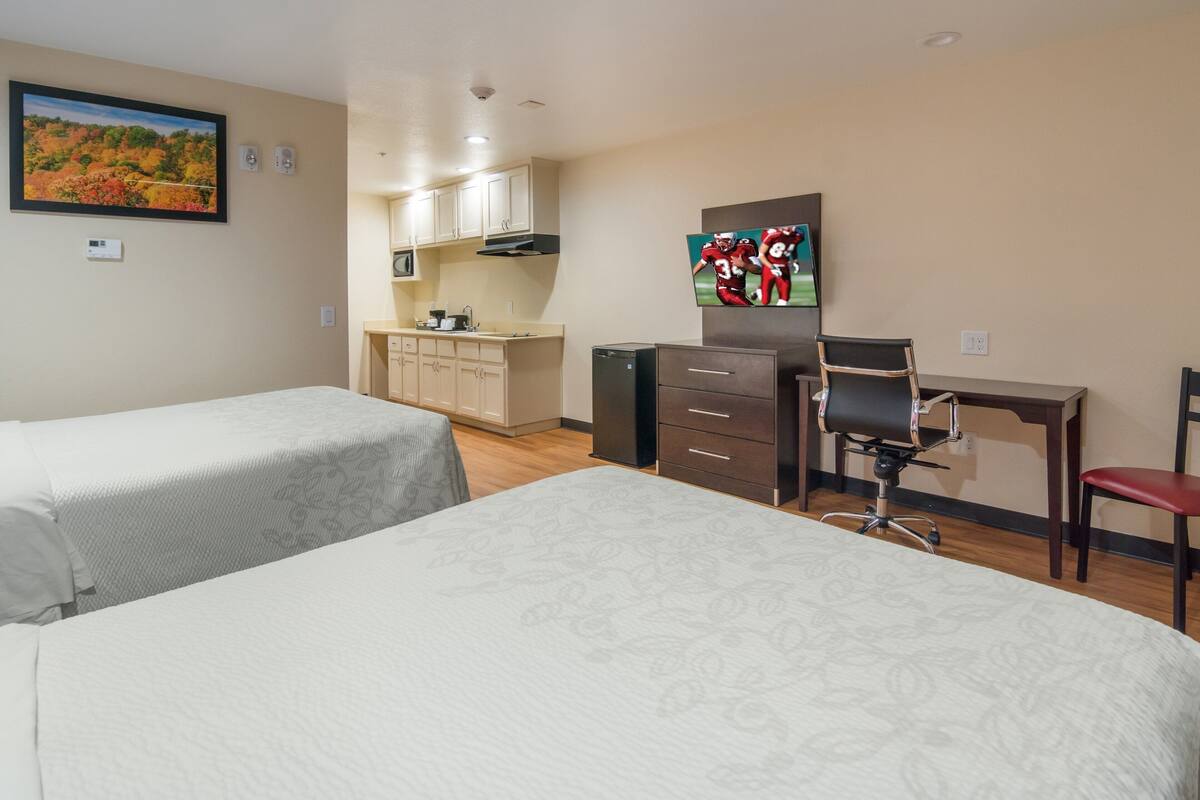 A hotel room with two beds, a desk, a chair, a TV, a kitchenette, and a painting on the wall.