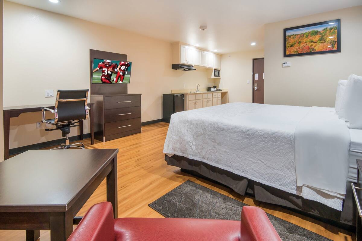 A hotel room with a bed, a desk, a chair, a TV, a kitchenette, and a painting on the wall.
