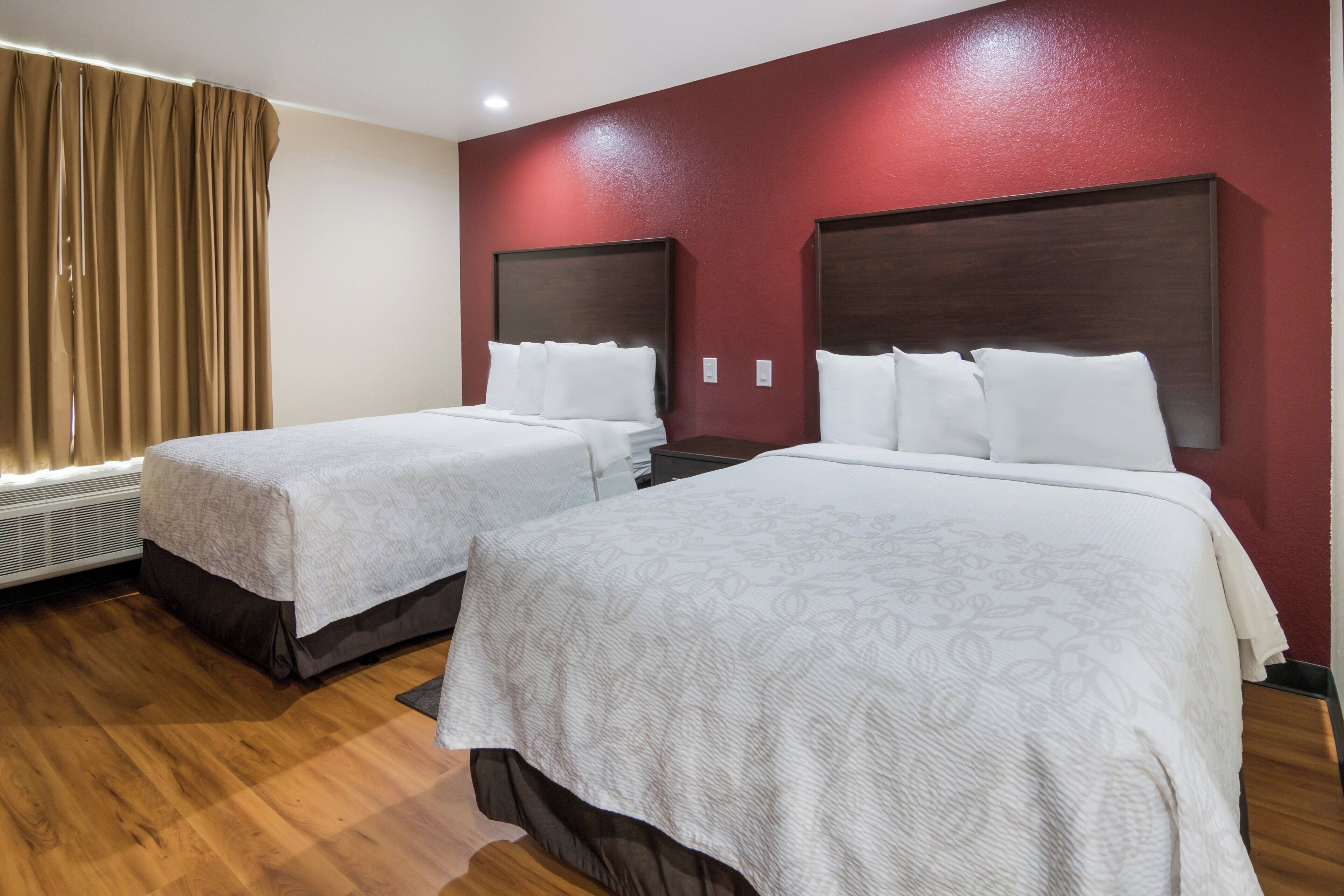 A hotel room with two beds, a red accent wall, and a window with curtains.
