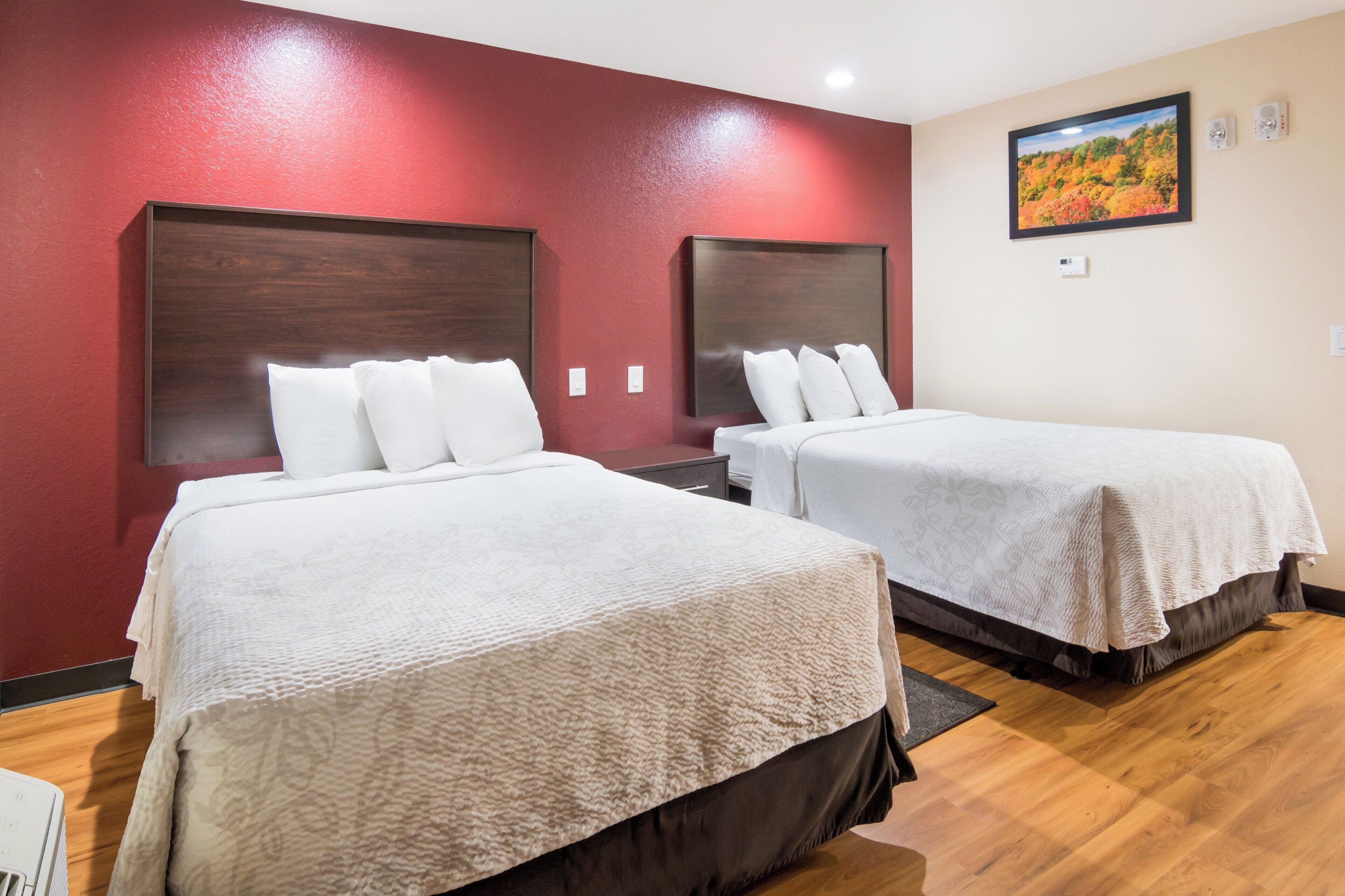 Habitación de hotel con dos camas, una pared de acento roja y un cuadro de un paisaje.