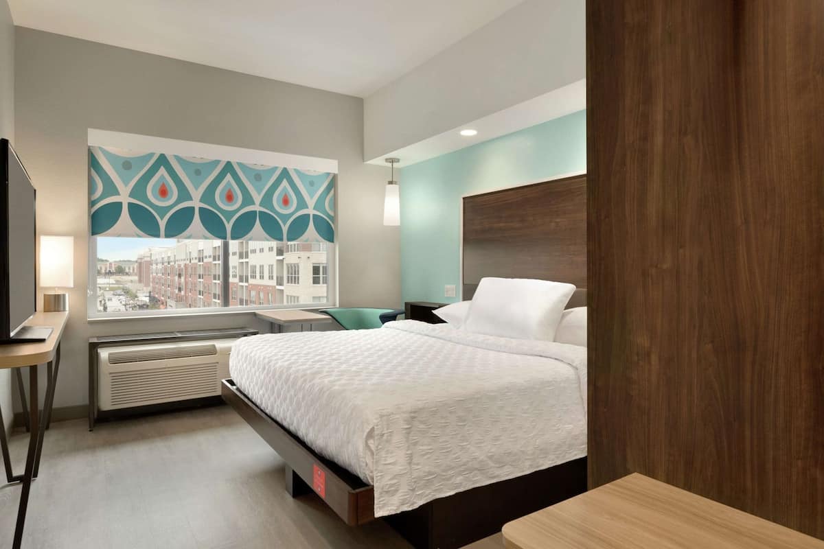 Habitación de hotel con cama, escritorio, silla, ventana con vistas a edificios y pared con diseño geométrico.