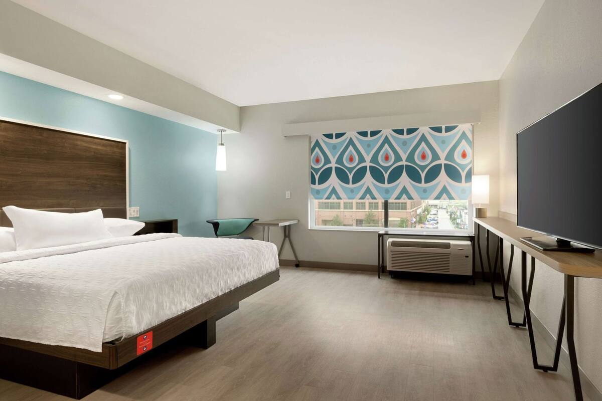 Habitación de hotel con cama, escritorio, silla, televisor y ventana con cortina estampada.