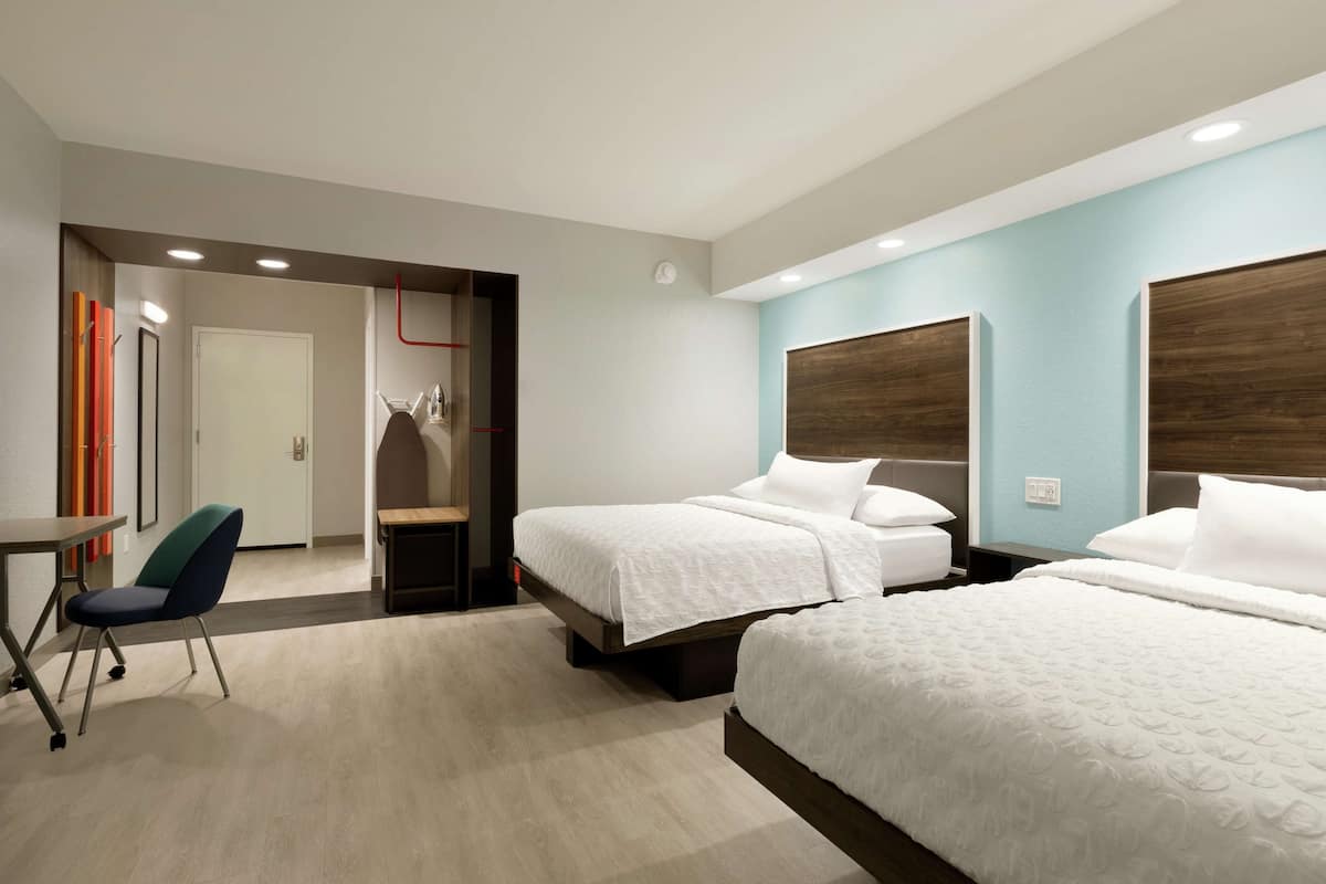 Habitación de hotel con dos camas, un escritorio y una silla. Se observa un perchero y una puerta que da a otra habitación.