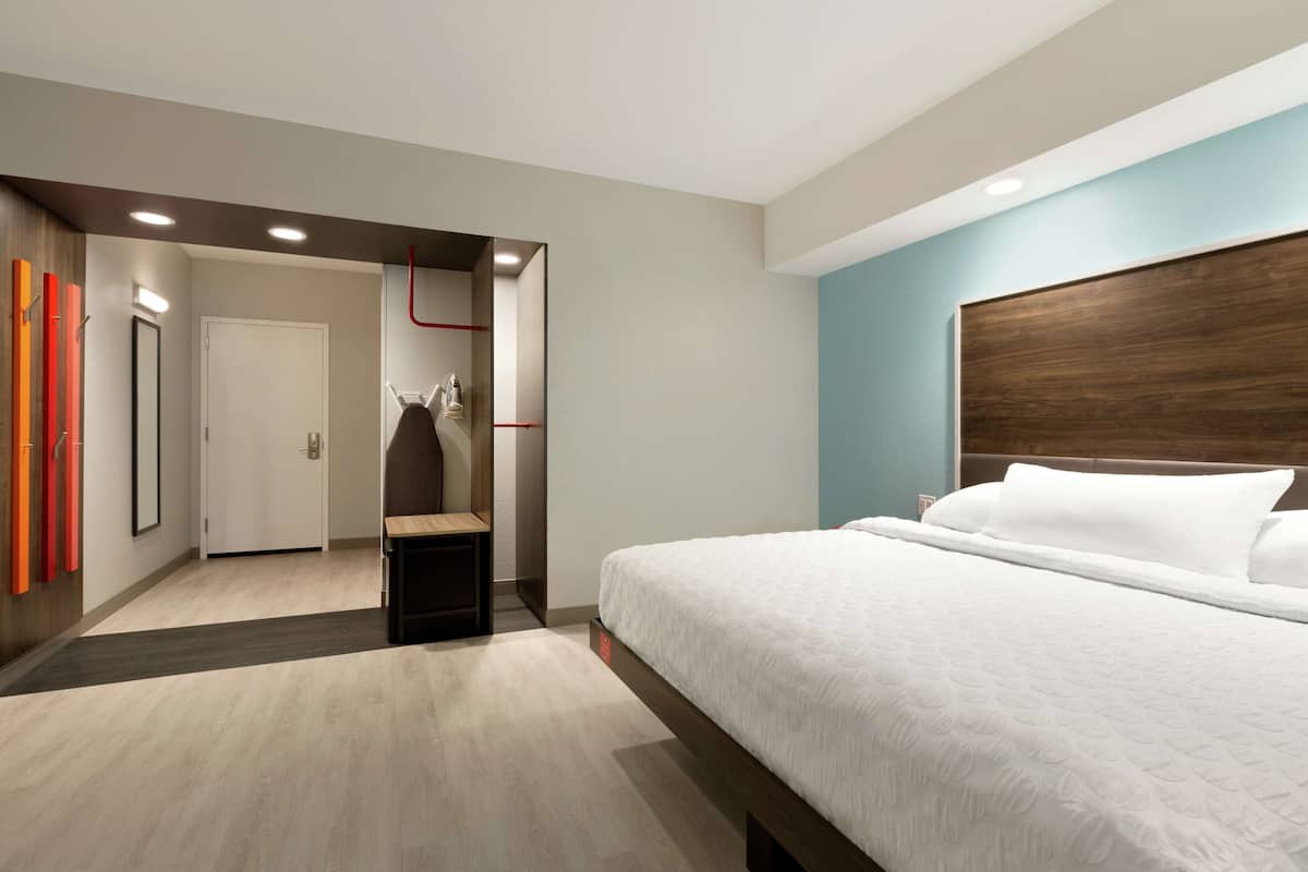 Habitación de hotel moderna con una cama grande, cabecera de madera y una puerta que da a un pasillo con espejo y detalles rojos.