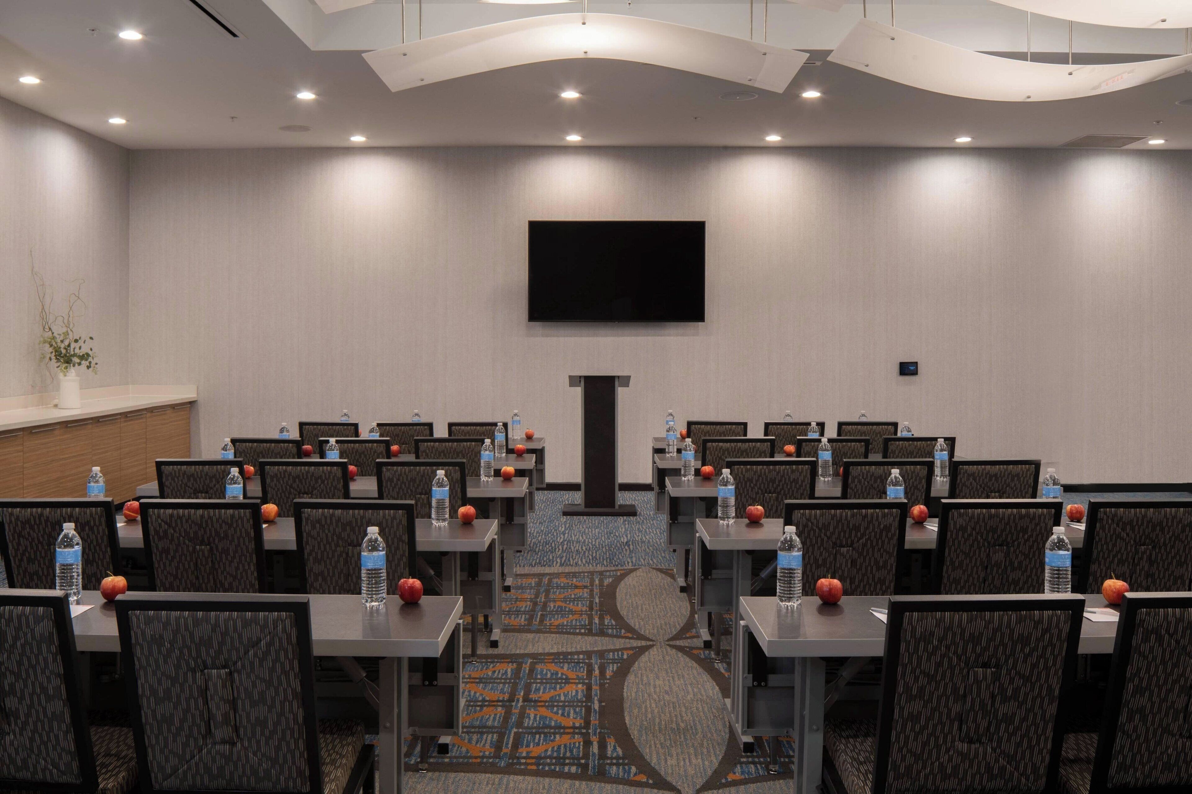 A conference room with rows of tables and chairs, equipped with water bottles and oranges, a flat-screen TV, and a podium.