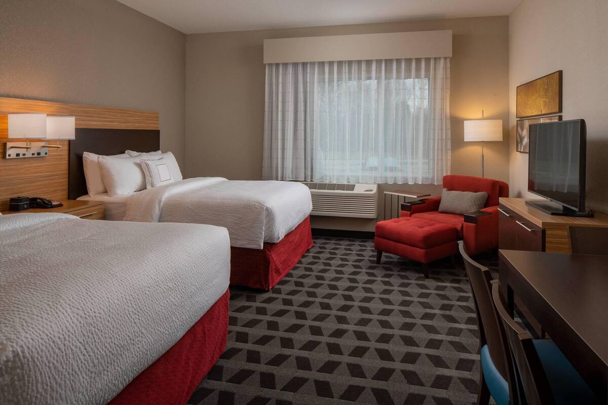 Ein Hotelzimmer mit zwei Betten, einem roten Sessel, einem Schreibtisch mit Fernseher und einem Fenster mit durchsichtigen Vorhängen.