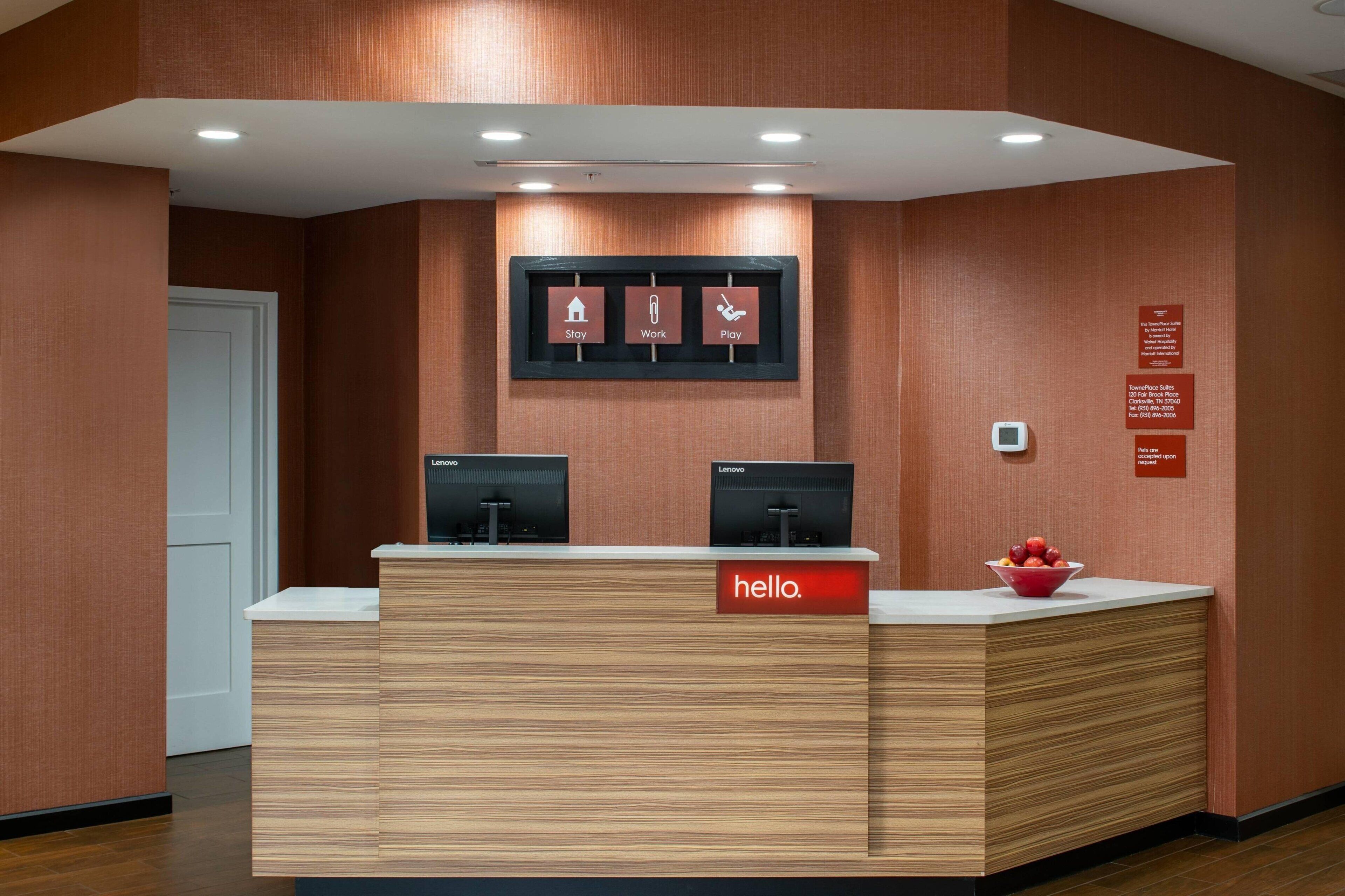 Un mostrador de recepción con dos monitores, un letrero que dice 'hello' y un cartel enmarcado con iconos para 'Stay', 'Work' y 'Play'.