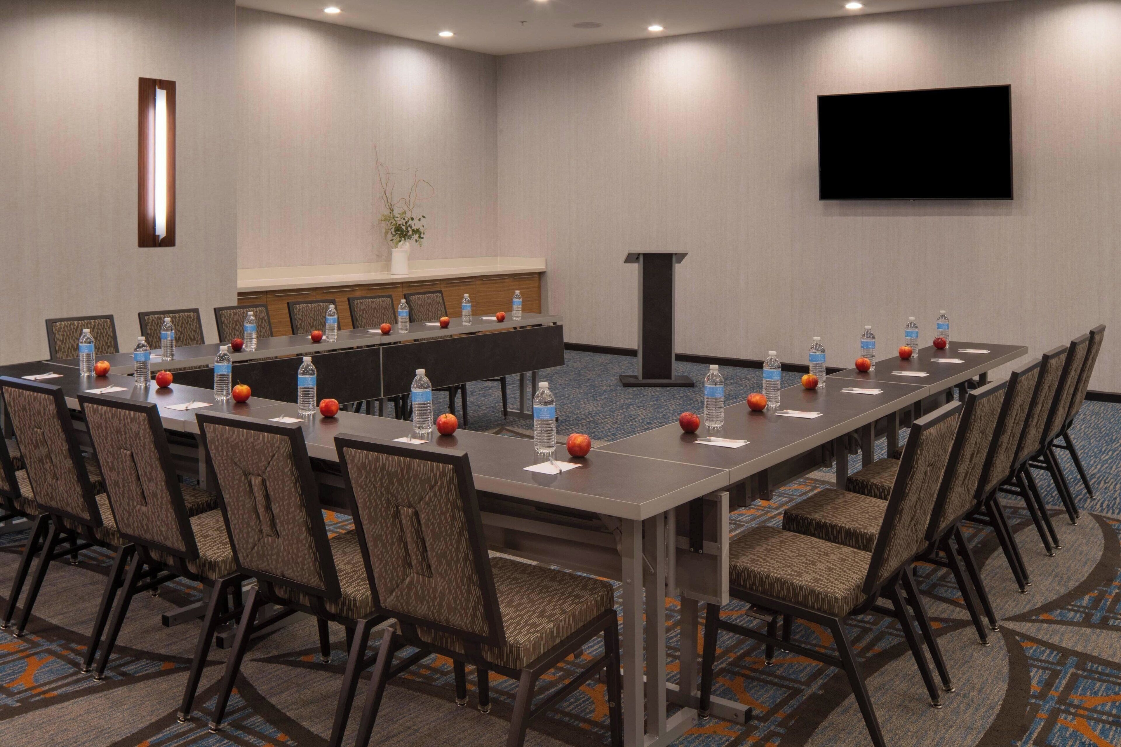 A conference room with a long table, chairs, water bottles, apples, and a TV screen.