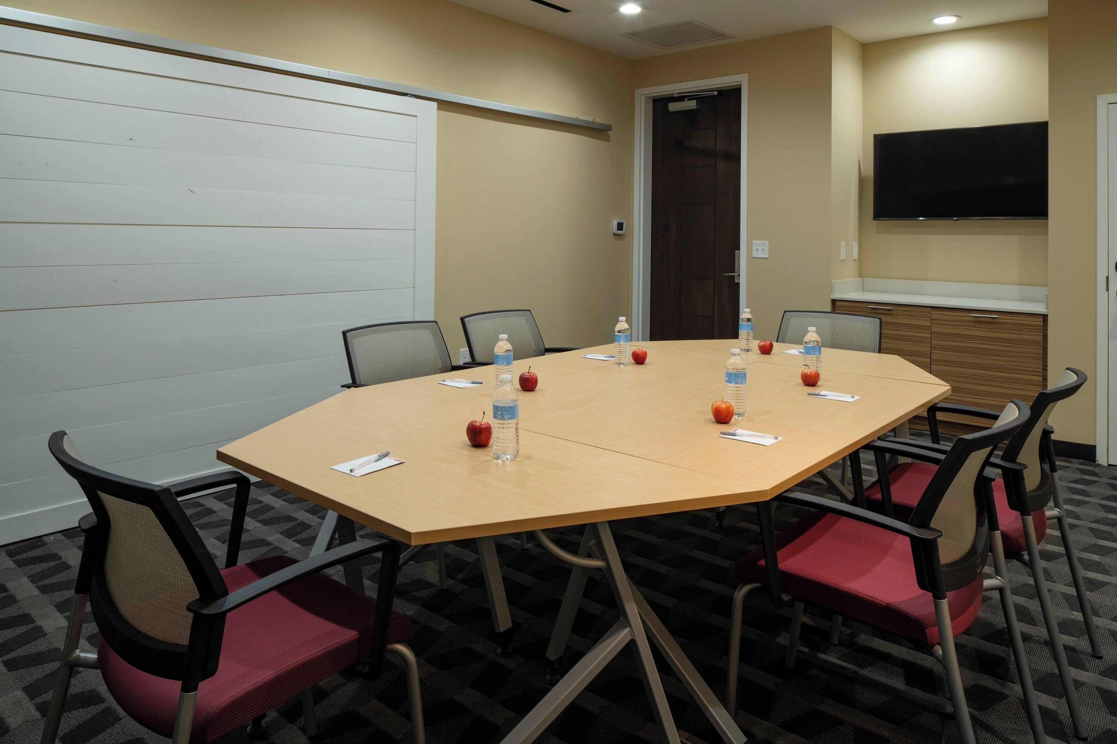 A conference room with a large table, chairs, water bottles, apples, and a TV on the wall.
