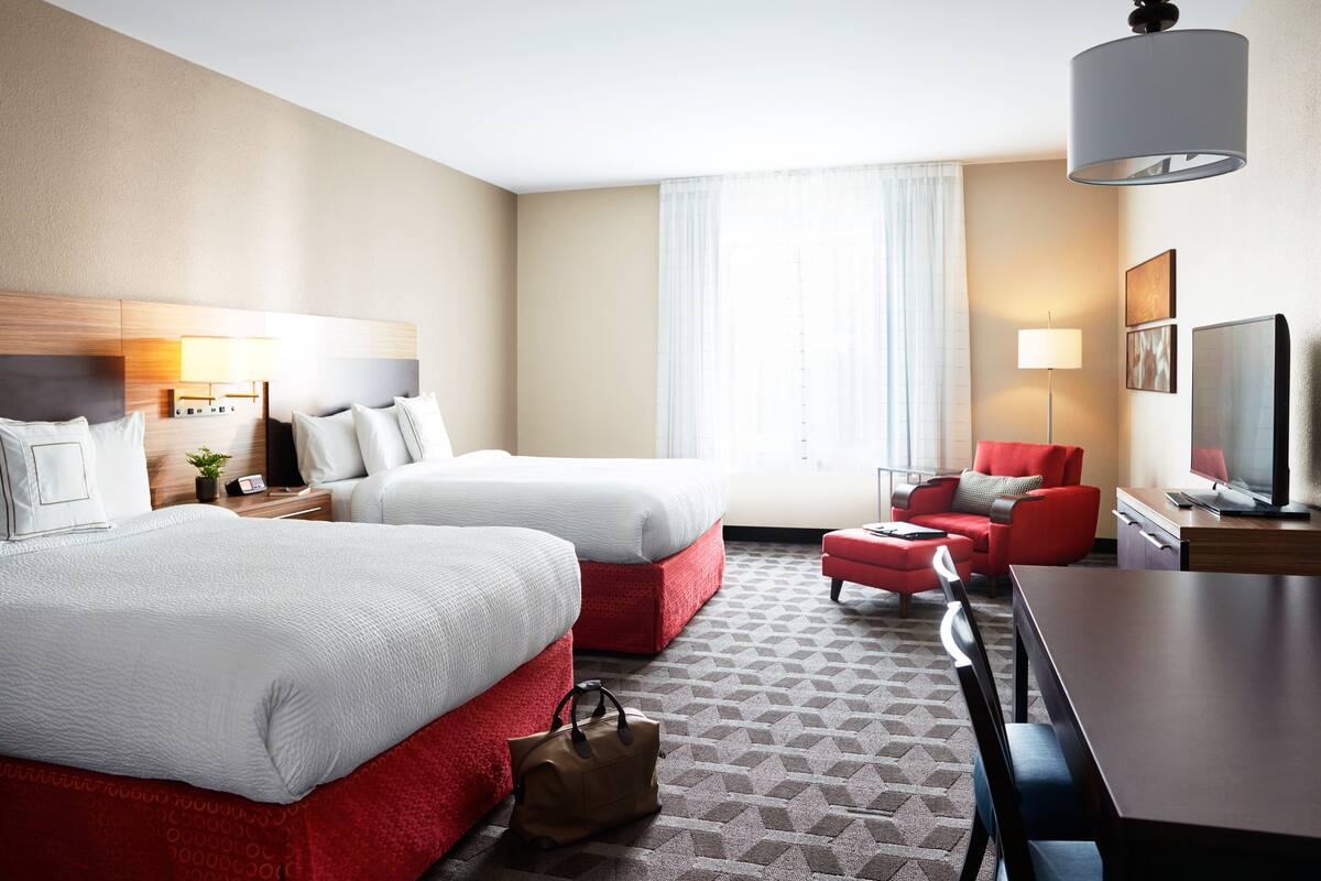 A hotel room with two beds, a desk, a chair, a TV, and a window with curtains.